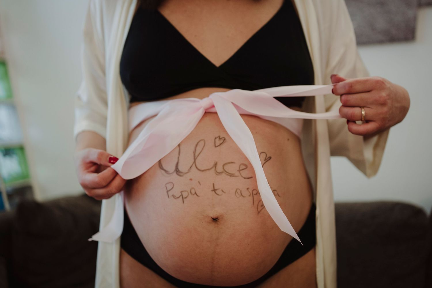 A pregnant belly with text written on it and a pink ribbon tied around.