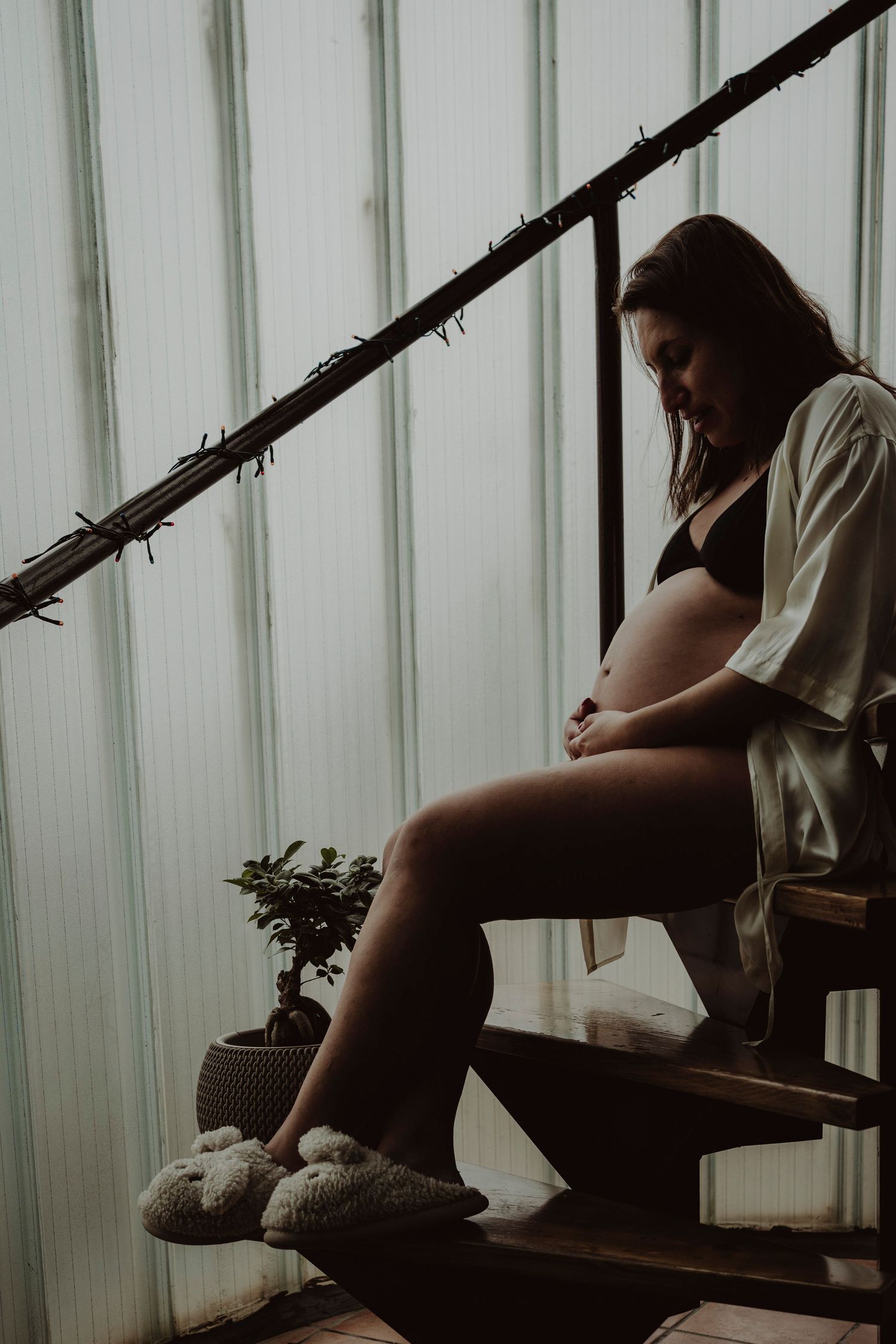 A silhouette of a pregnant person sitting on wooden stairs wearing fuzzy slippers.
