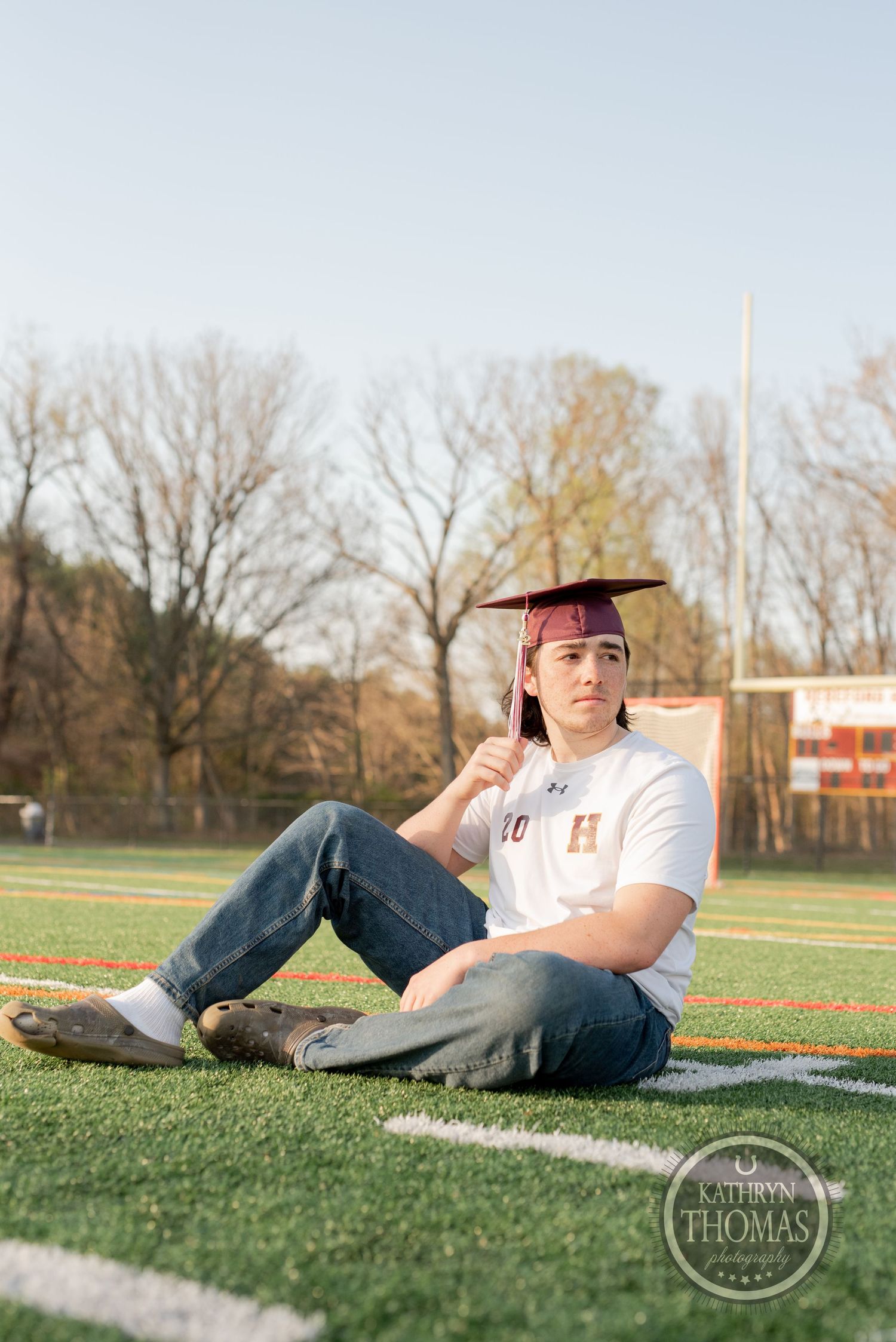 Graduate in casual attire and cap sits thoughtfully on sports field during golden hour.