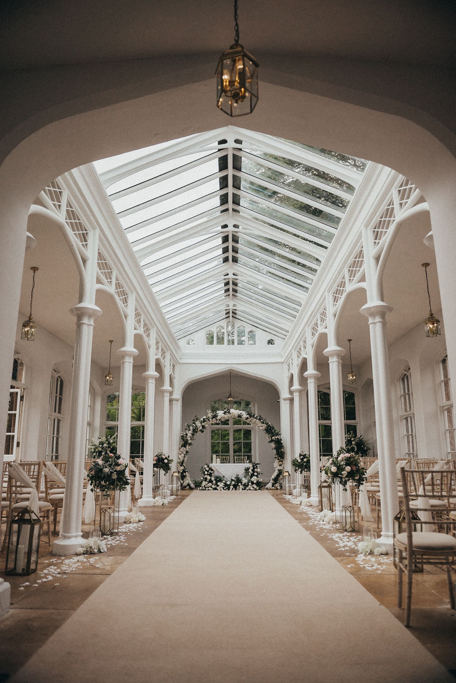 Elegant wedding aisle with glass ceiling, white columns and scattered petals leading to floral arch.