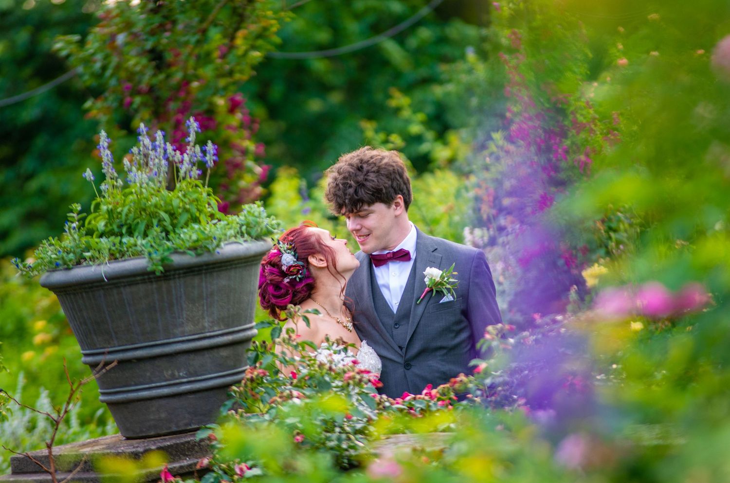 Romantic garden wedding portrait surrounded by purple flowers and lush greenery.