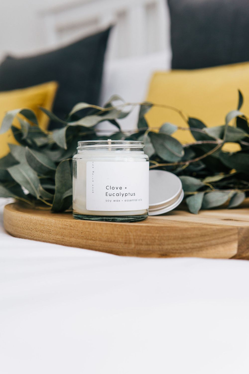 White scented candle jar surrounded by green eucalyptus leaves on wooden serving board.
