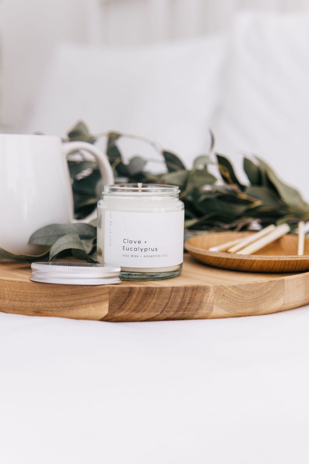 Minimalist scented candle on wooden tray with eucalyptus leaves and white ceramic mug in background.