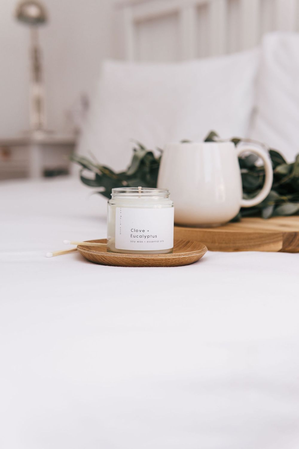 White candle jar on wooden coaster with greenery and ceramic mug in soft lighting.