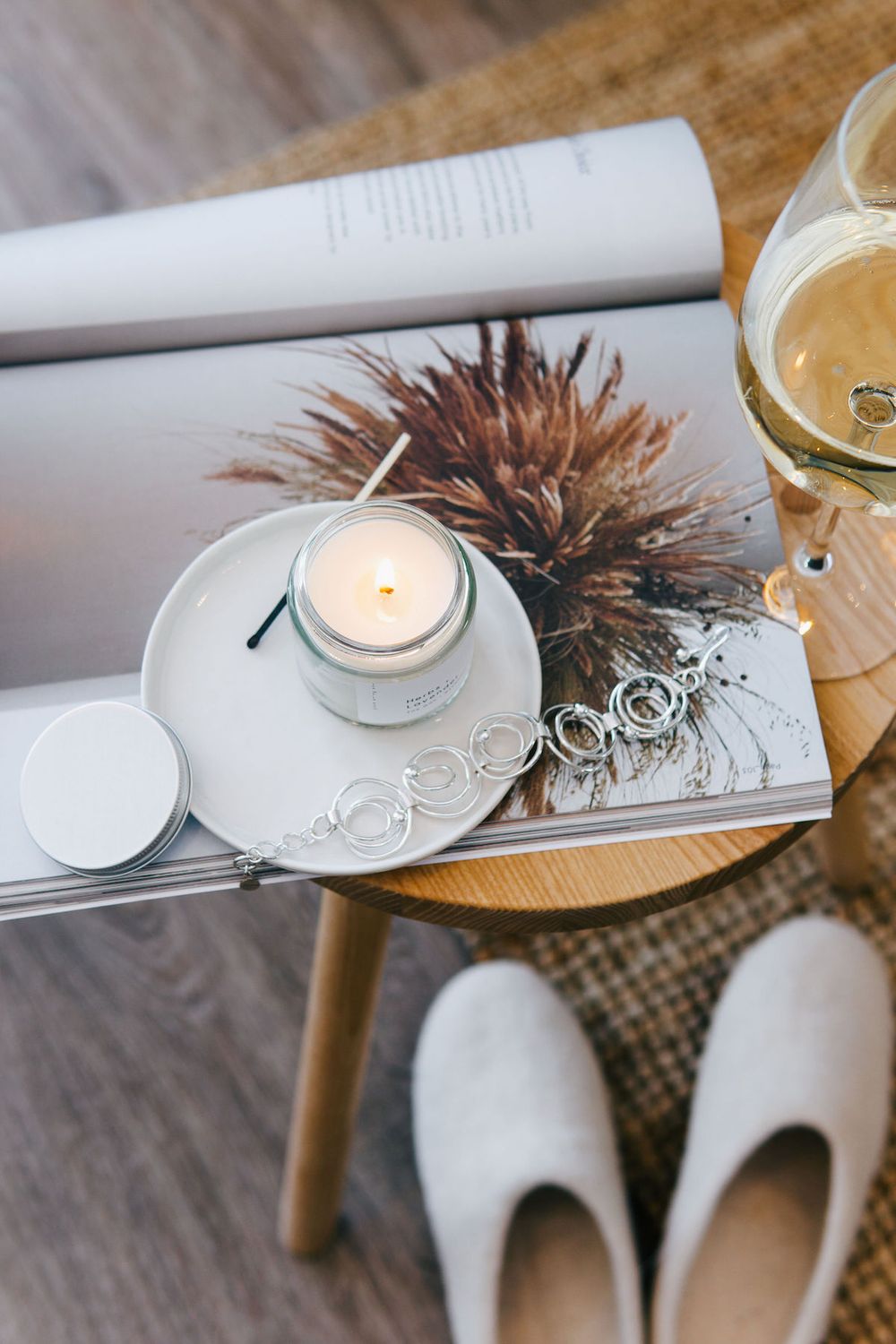 Lit candle on white plate with dried flower arrangement and wine glass on wooden table.