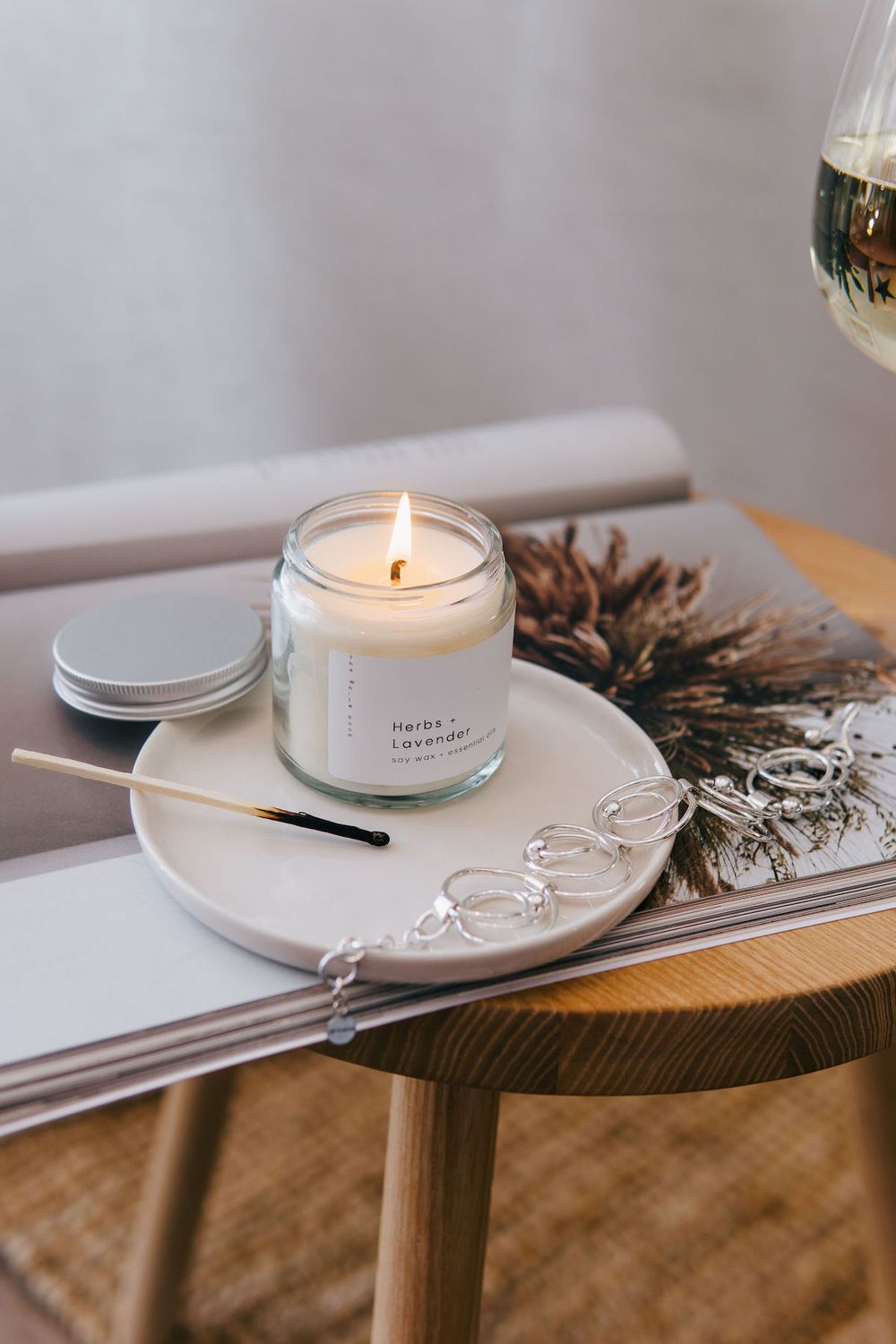 Burning candle in glass jar on white ceramic plate with dried botanical arrangement.
