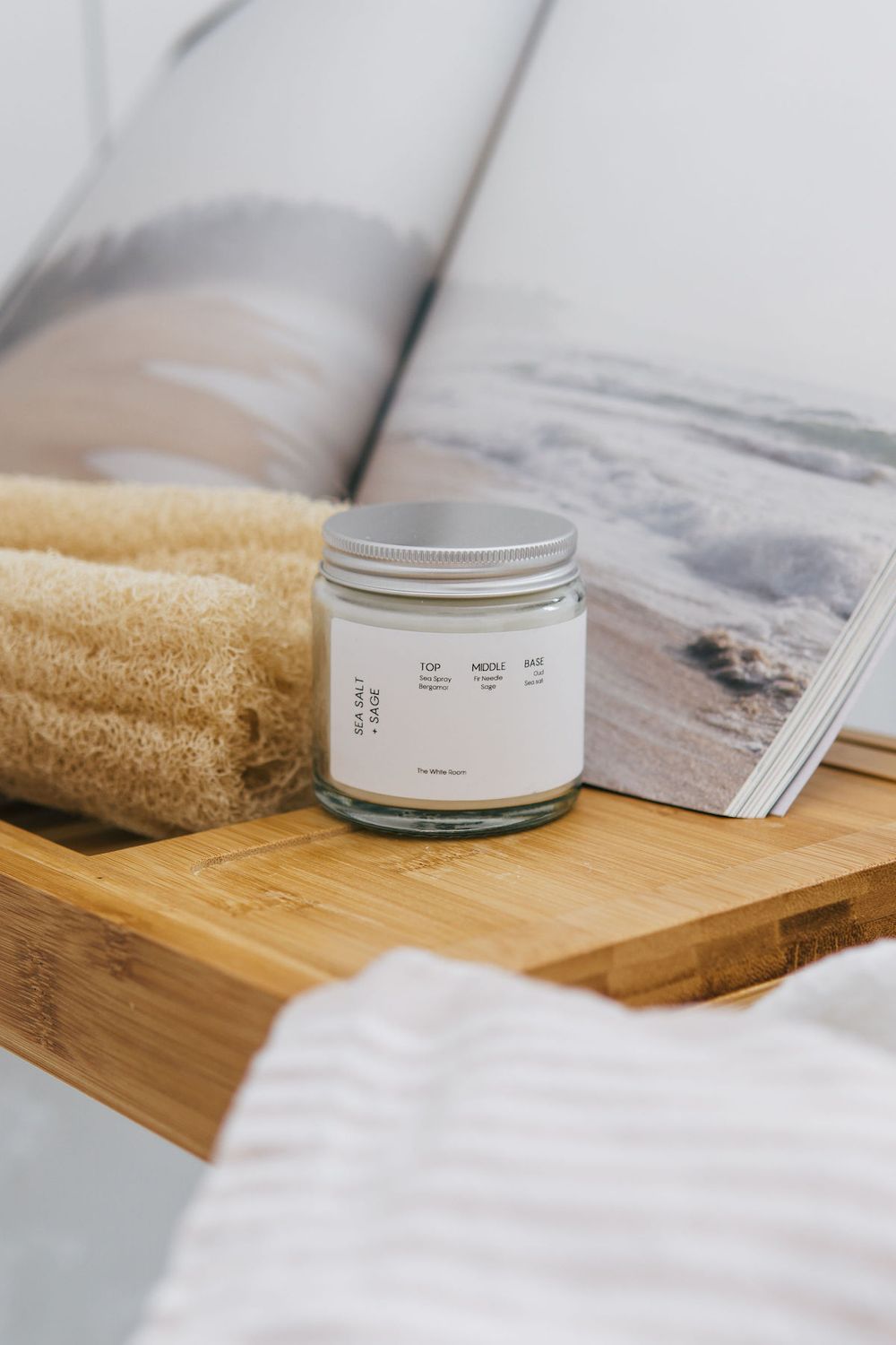 Glass jar candle on wooden bath tray with beige towel and ocean photography book.