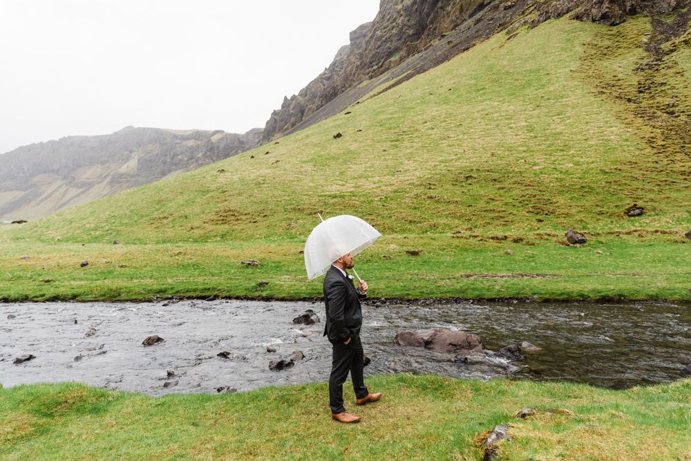 Person holding white umbrella walks along stream in misty mountainous landscape.