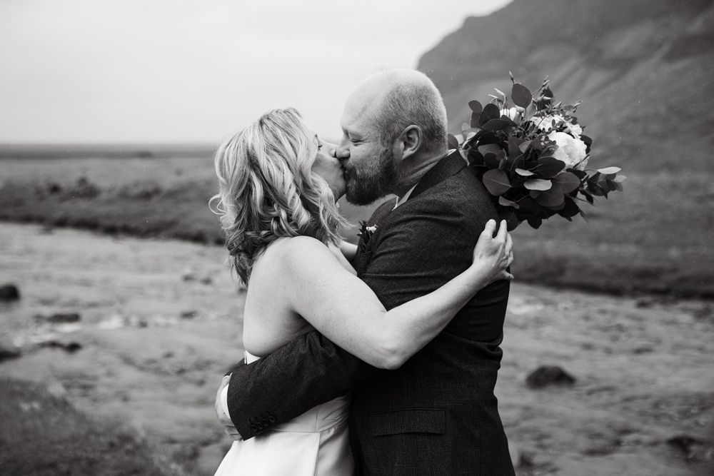 A romantic wedding kiss captured in black and white against a dramatic landscape.