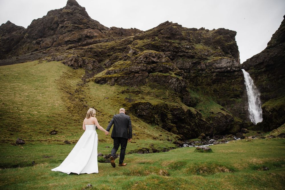 A couple walks toward a dramatic waterfall cascading down moss-covered cliffs in the rugged highlands of Iceland.