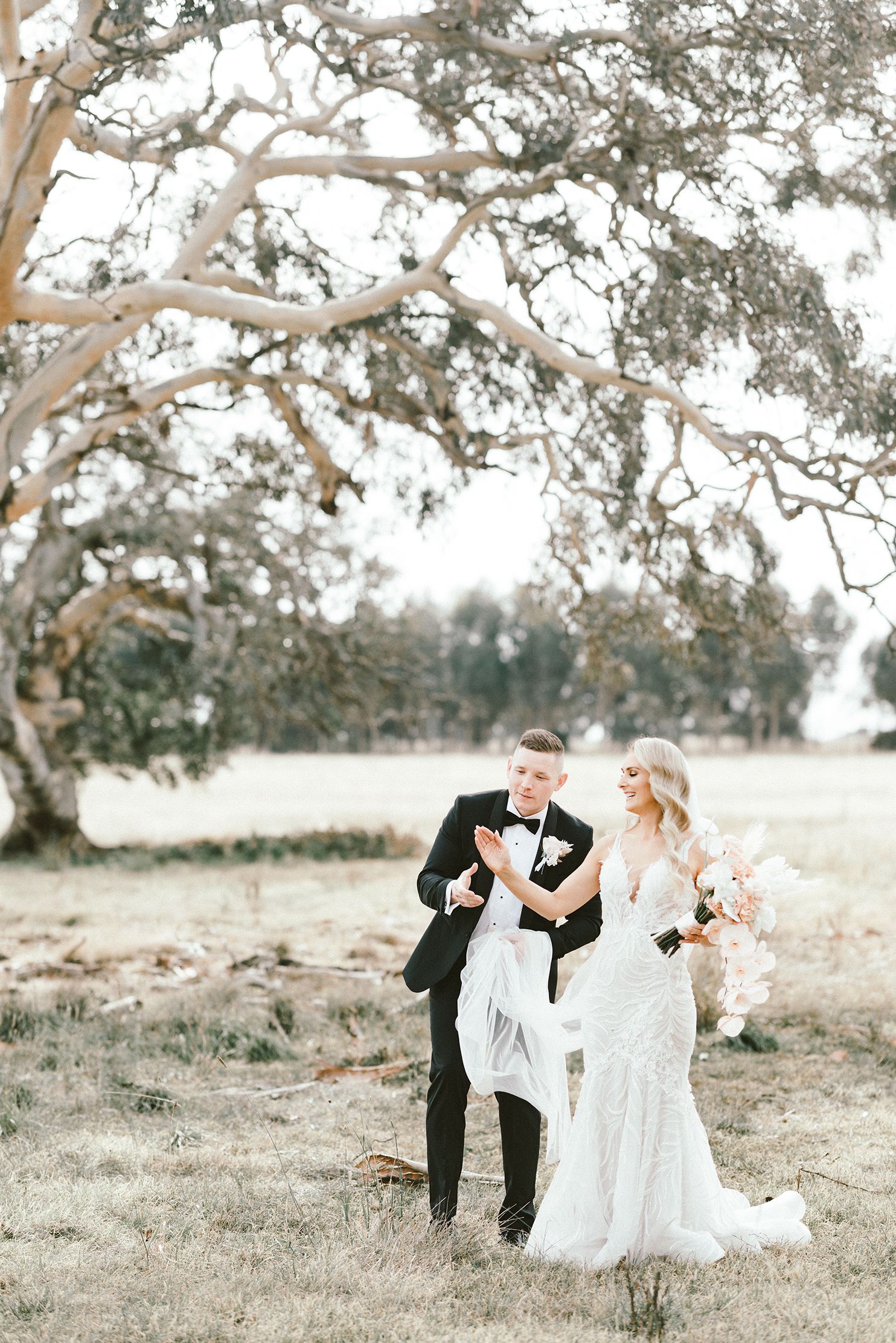 Newlyweds share a joyful moment under a grand tree in an open field.