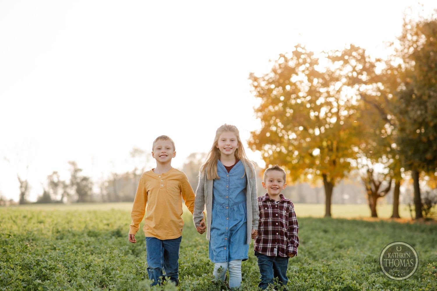 Three children standing together in a green field during sunset with golden light filtering through trees.