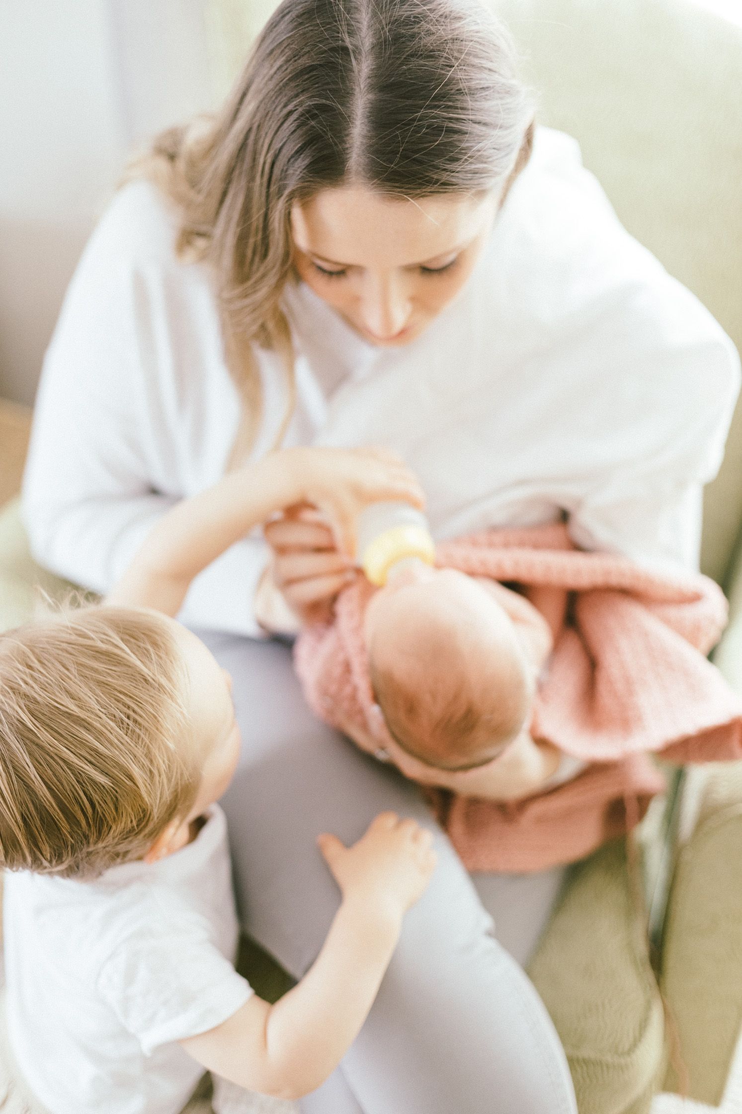 Family moment as toddler reaches up while caregiver feeds infant in cozy living room setting.