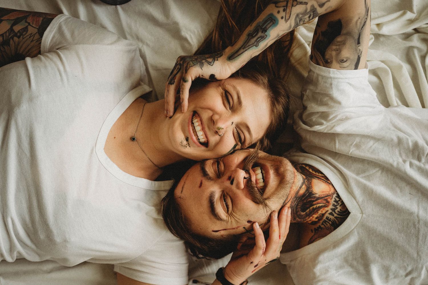 Close-up portrait of a joyful couple laying on white bedding with visible tattoos.