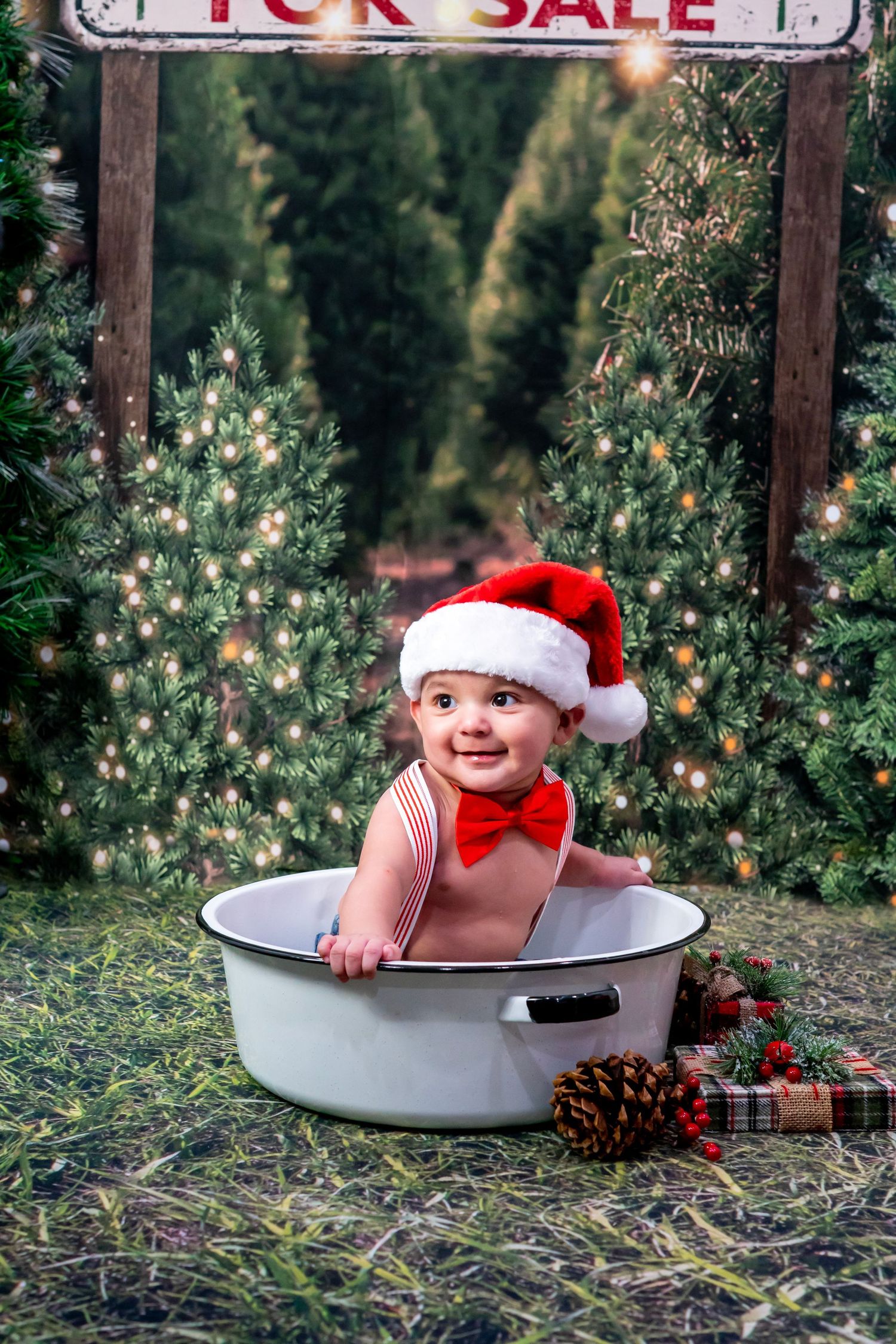 Smiling baby in Santa hat and red bow tie sitting in white metal tub at Christmas tree farm.