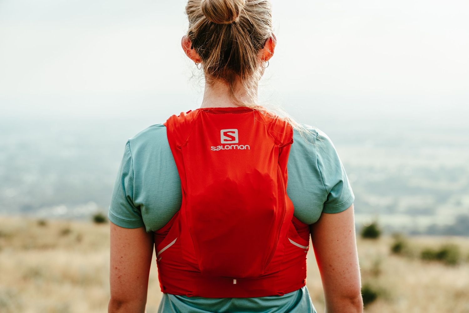 Hiker wearing a red Salomon hydration vest and teal shirt stands on a hilltop overlooking a misty valley.