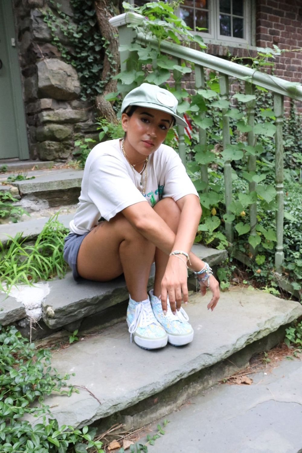 Young woman in mint cap, graphic tee and white sneakers sits on stone steps surrounded by lush green ivy.