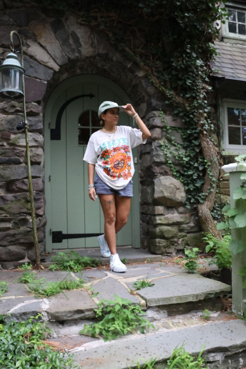 Stylish woman in oversized graphic tee, denim shorts and mint cap stands by a green arched door of a stone building.