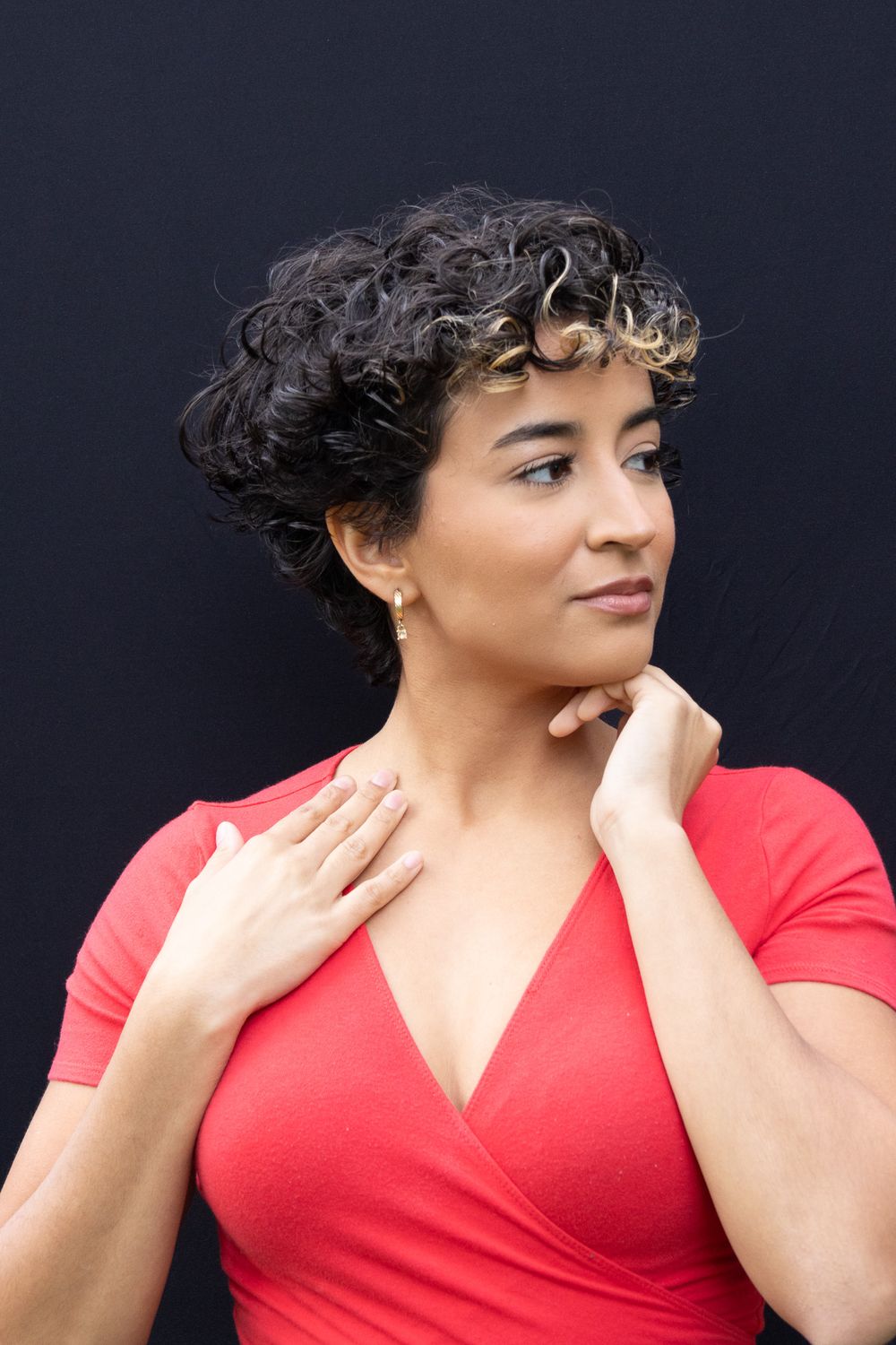Young woman with short curly hair in a red wrap top posing against a dark black wall.