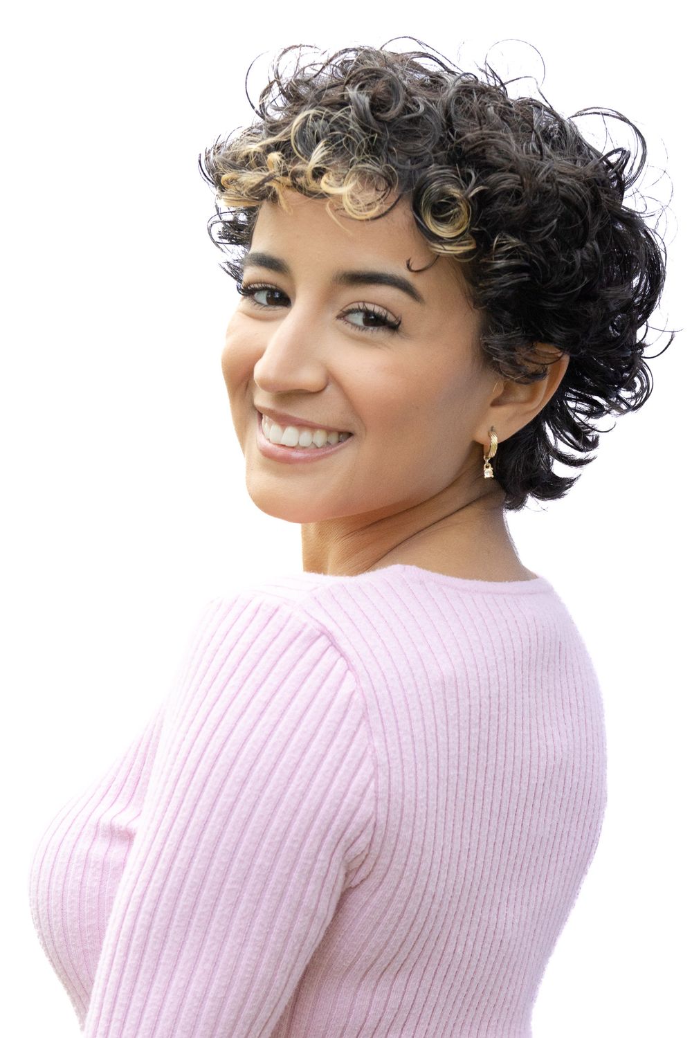 Smiling young woman with short curly highlighted hair wearing a pink ribbed sweater against white background.