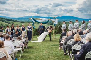 Wedding guests seated in white chairs on a grassy hilltop during an outdoor ceremony with mountain backdrop.