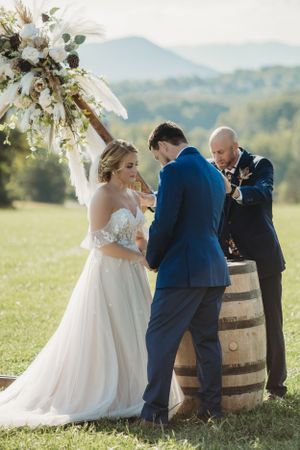 Wedding ceremony taking place beside wooden barrels and floral arrangements in a grassy field.