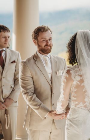 Wedding party members in beige suits and white dress stand together against a scenic backdrop.