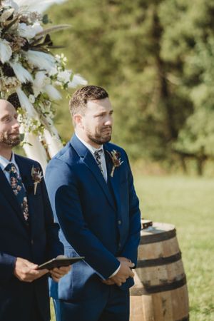 Wedding party members in blue suits gathered around rustic wooden barrels during ceremony.