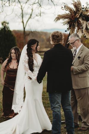 Wedding ceremony in progress with guests and wedding party in formal attire against autumn backdrop.