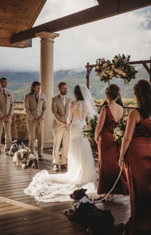 Wedding ceremony on covered deck with burgundy bridesmaids dresses and beige groomsmen suits.