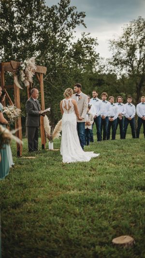 Wedding ceremony taking place on a grassy hillside with wooden arch and mountain backdrop.