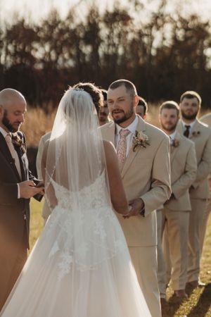 Bride and groom share an intimate moment during their outdoor autumn wedding ceremony with wedding party in beige suits.
