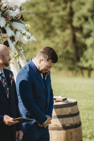 Groomsmen in navy suits standing near wooden barrels at an outdoor wedding venue.