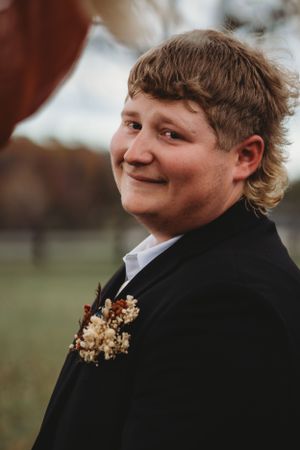 Groomsman in black suit with dried flower boutonniere smiles during outdoor wedding ceremony.