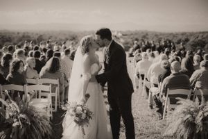Black and white panoramic shot of a rustic outdoor wedding ceremony with guests seated on chairs.