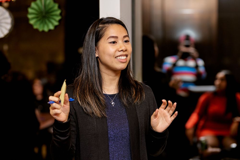 Speaker gestures animatedly while presenting at an indoor event with decorative paper fans on the wall.