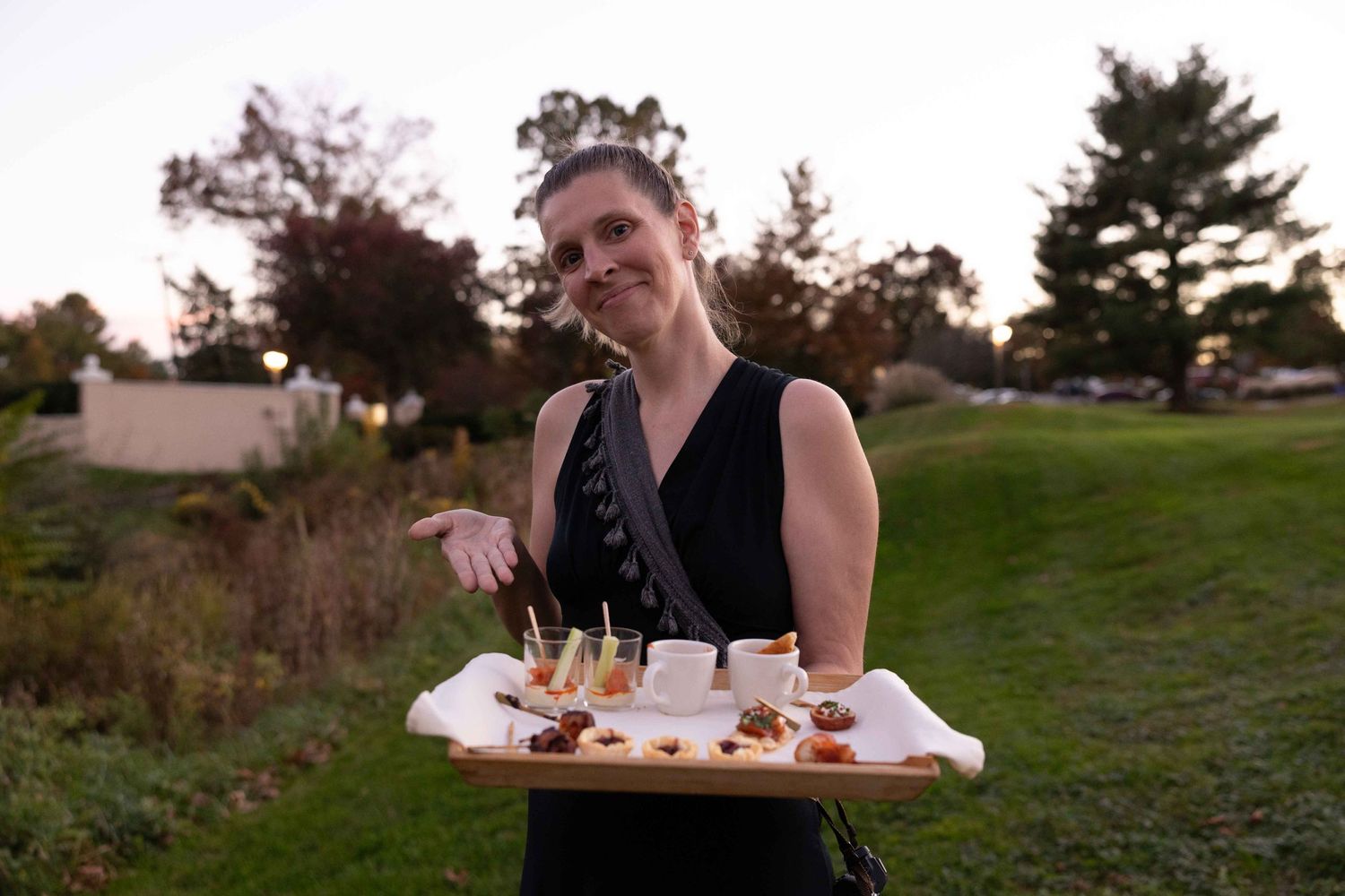 A server holds a wooden tray of appetizers at an outdoor event during sunset.