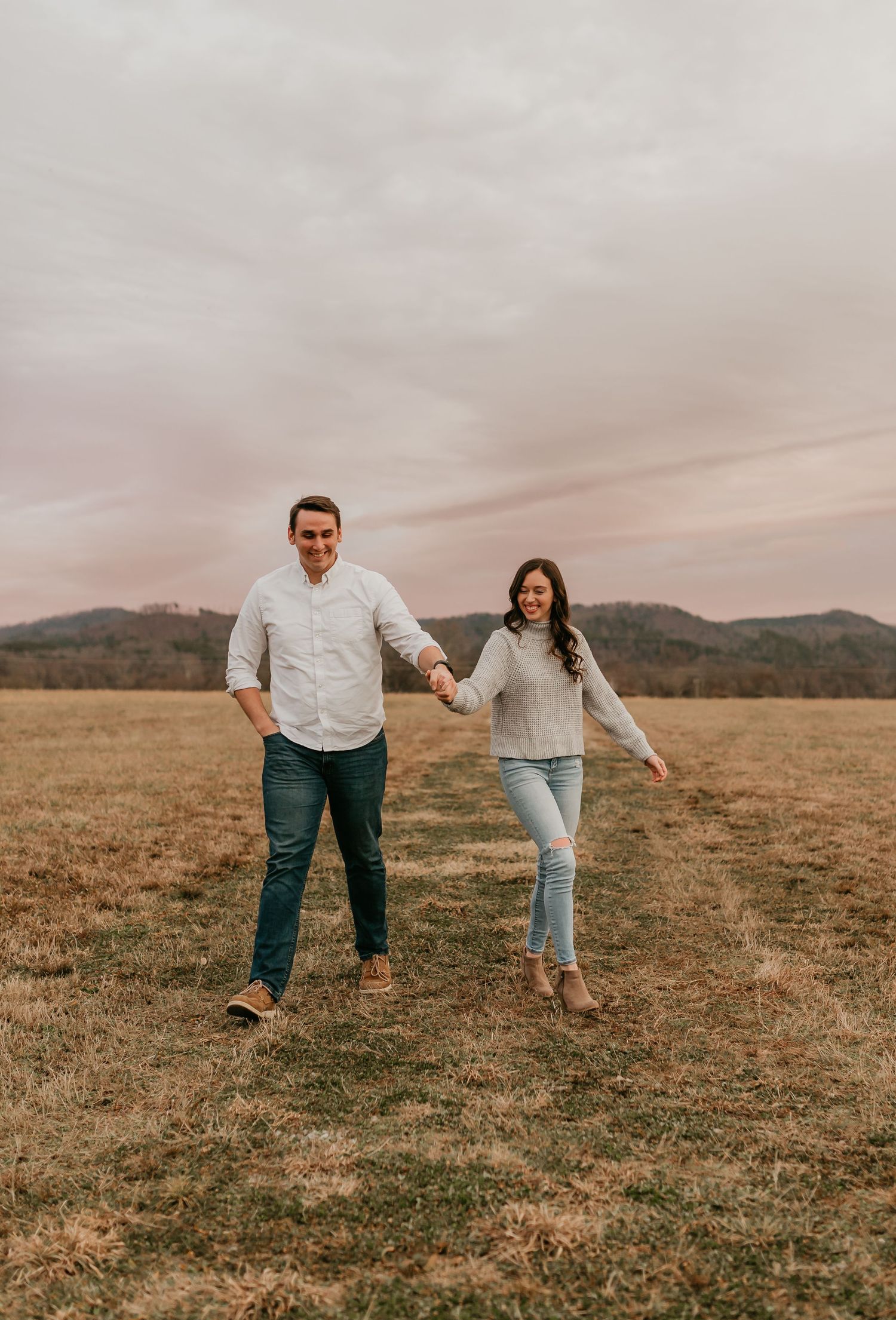 Happy couple holding hands and walking through an open field with mountains and cloudy sky.