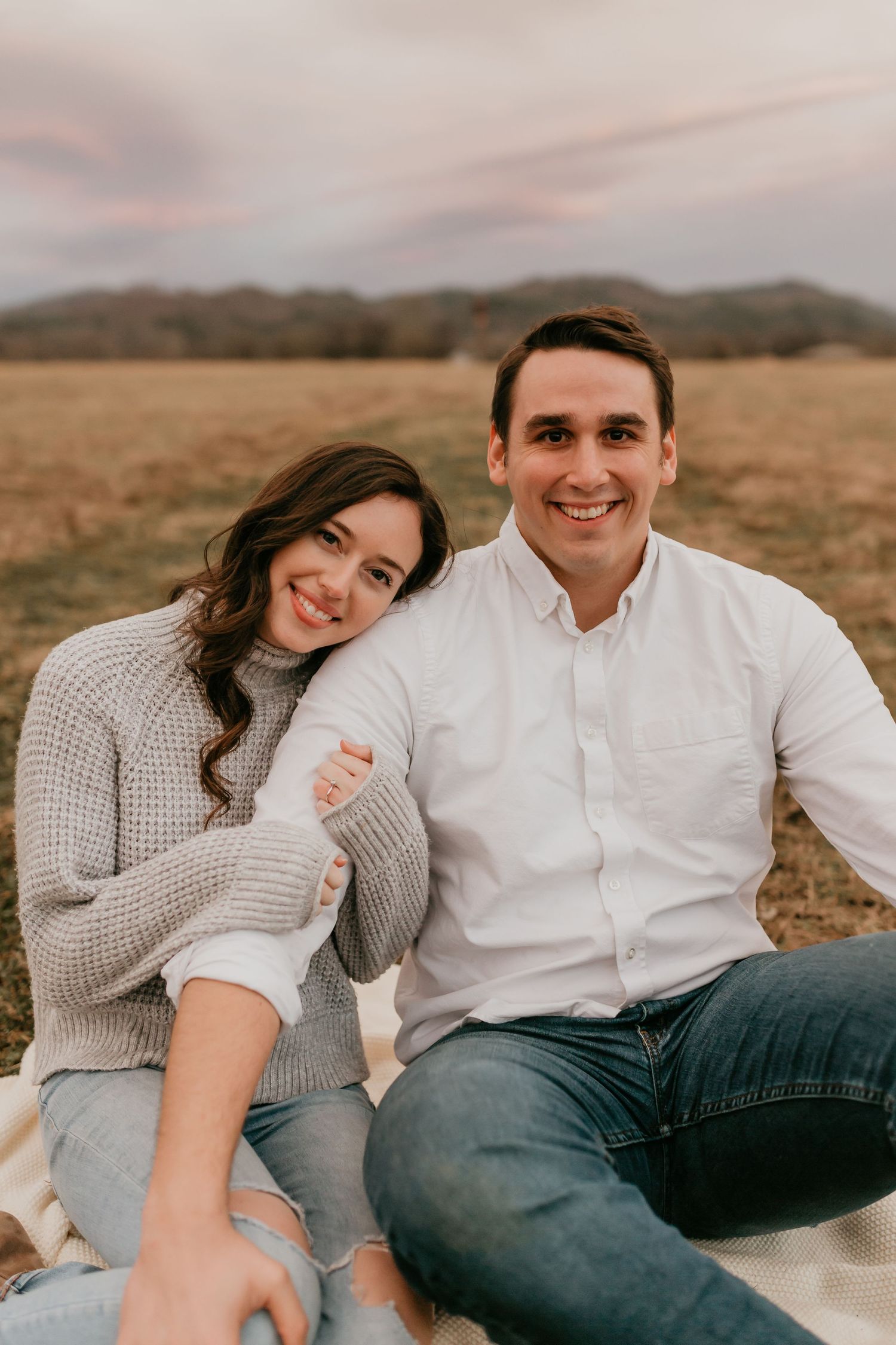 Smiling couple sitting together on a blanket in a field, woman leaning on mans shoulder.