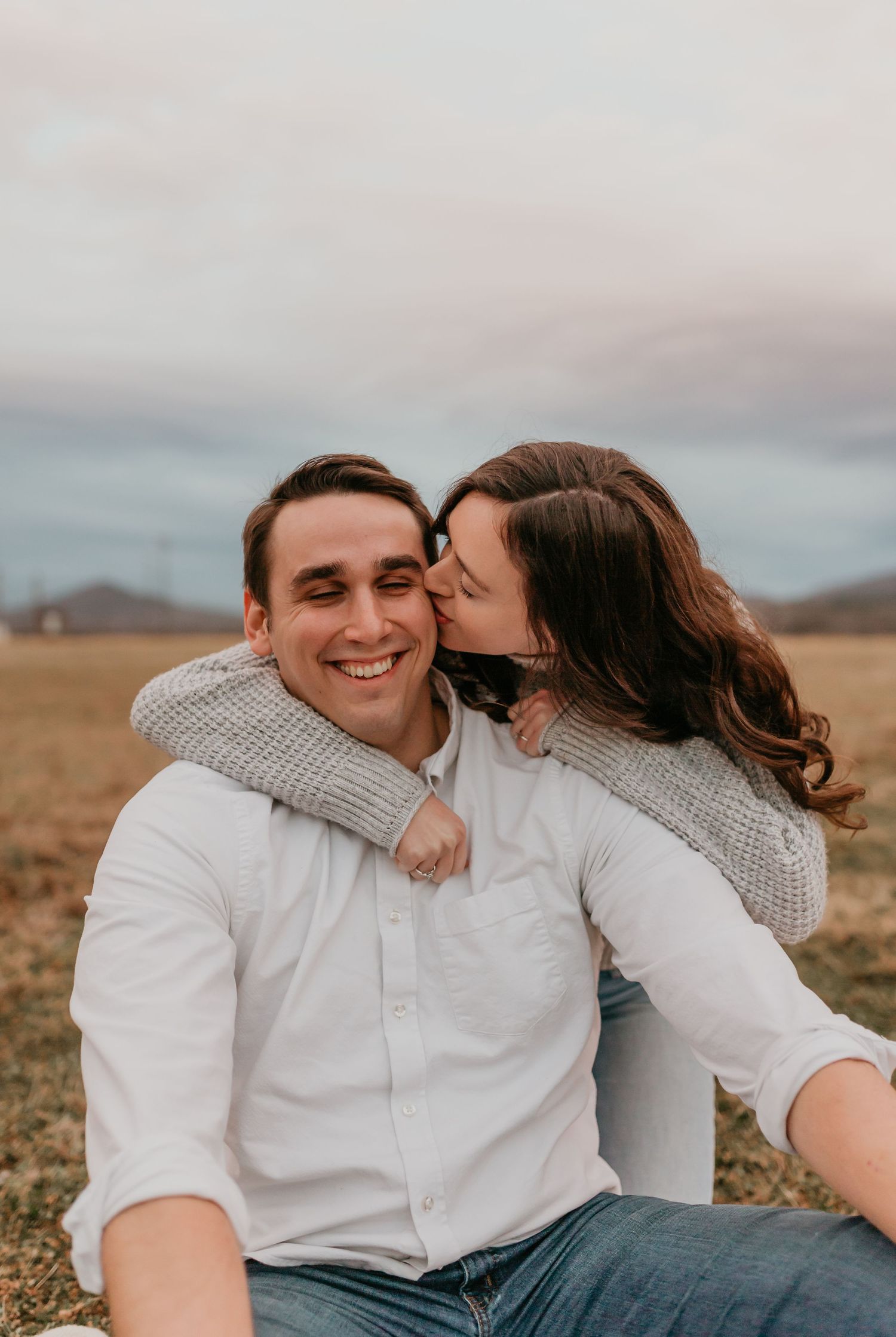 Woman kissing man on the cheek as he smiles broadly while sitting in a golden field.