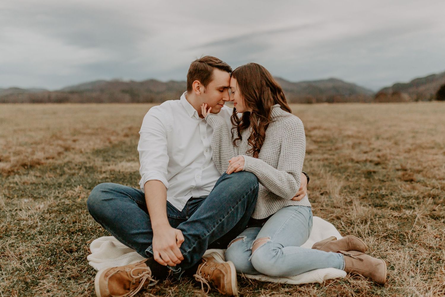 Couple sitting cross-legged in a field, leaning in close for a kiss with mountains behind.