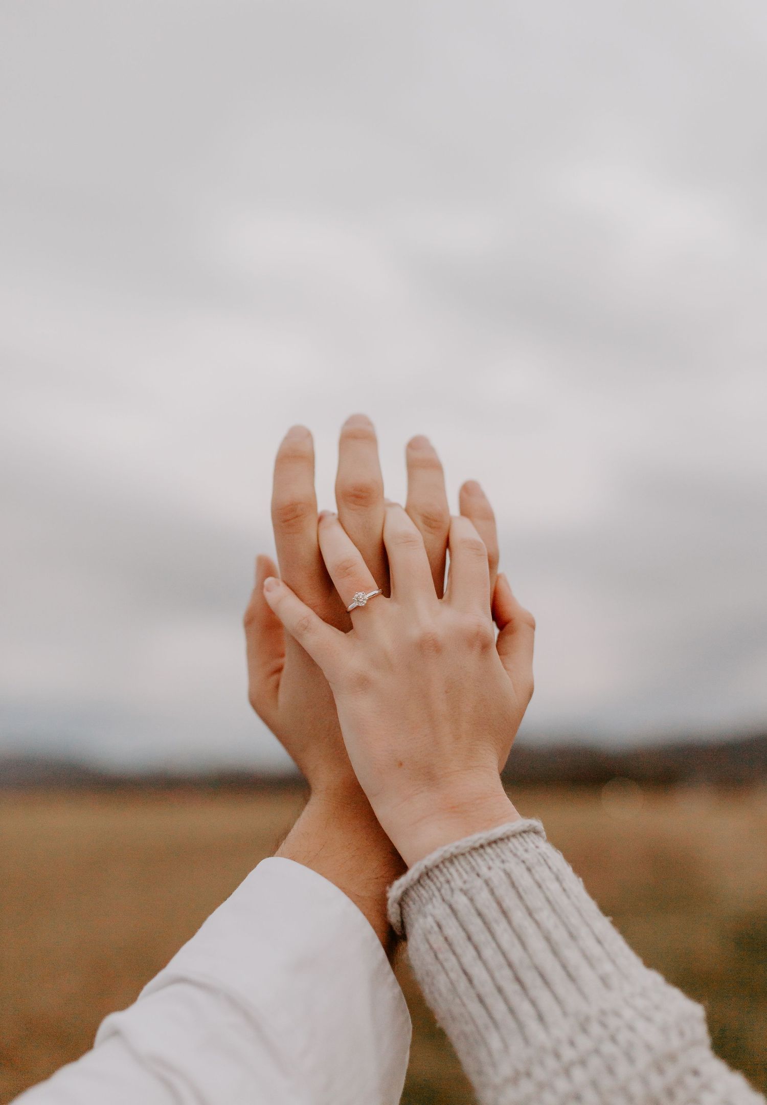 Engaged couple holding hands up toward cloudy sky, showing off engagement ring in a field.