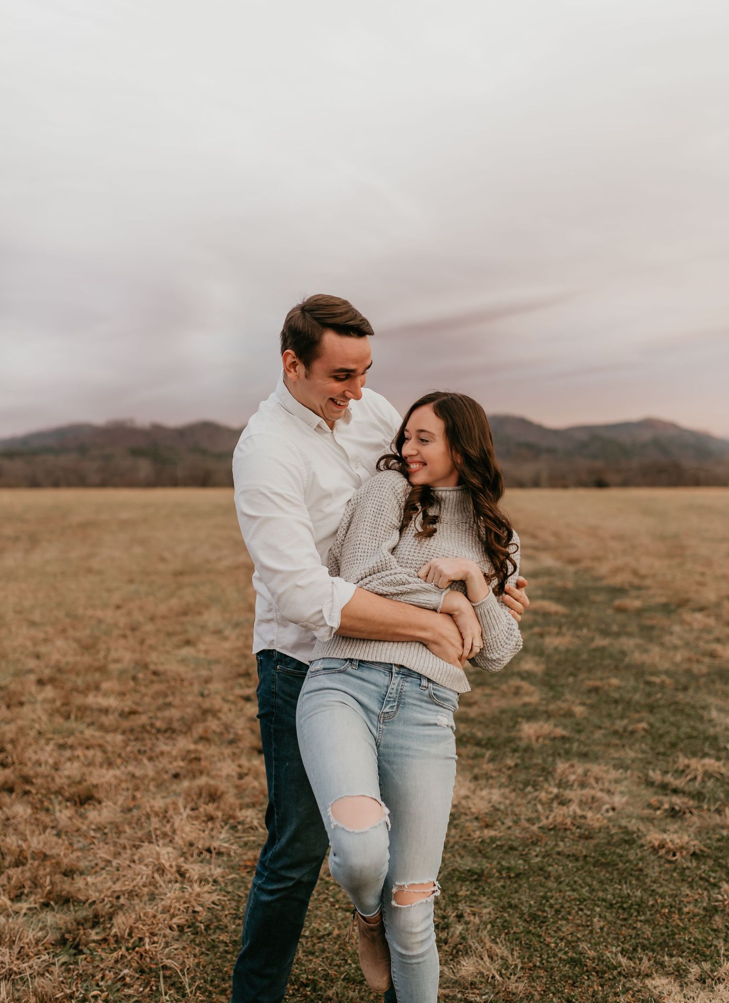 Man lifting and embracing woman in a field with mountains in the background during golden hour.