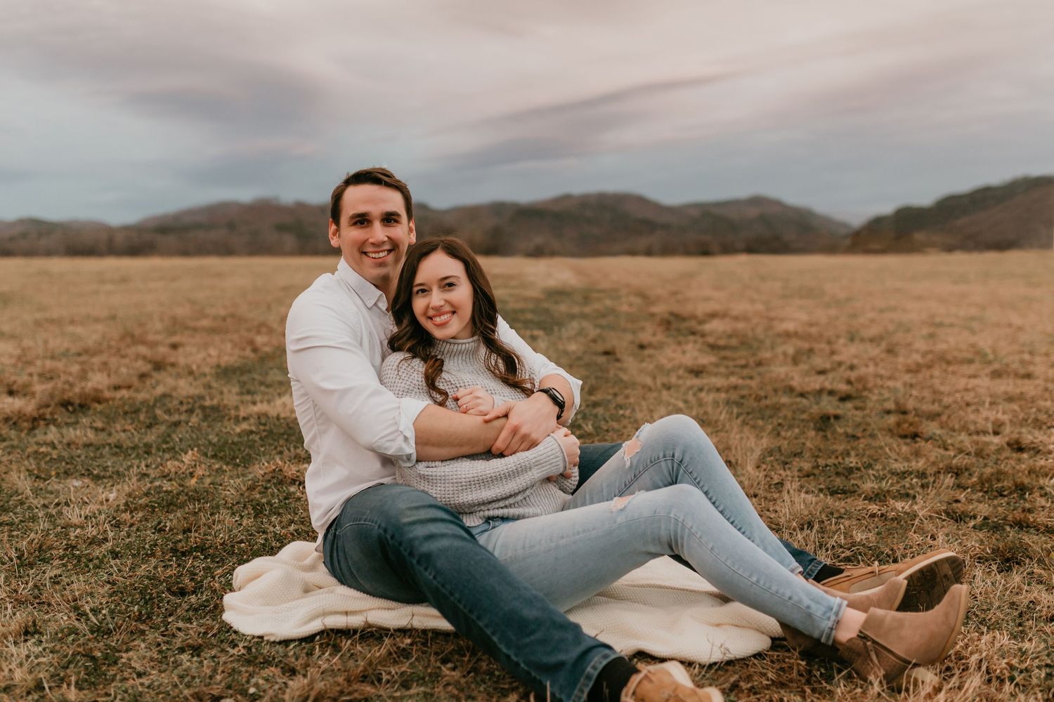 Happy couple sitting on a blanket in an open field, smiling at the camera together.