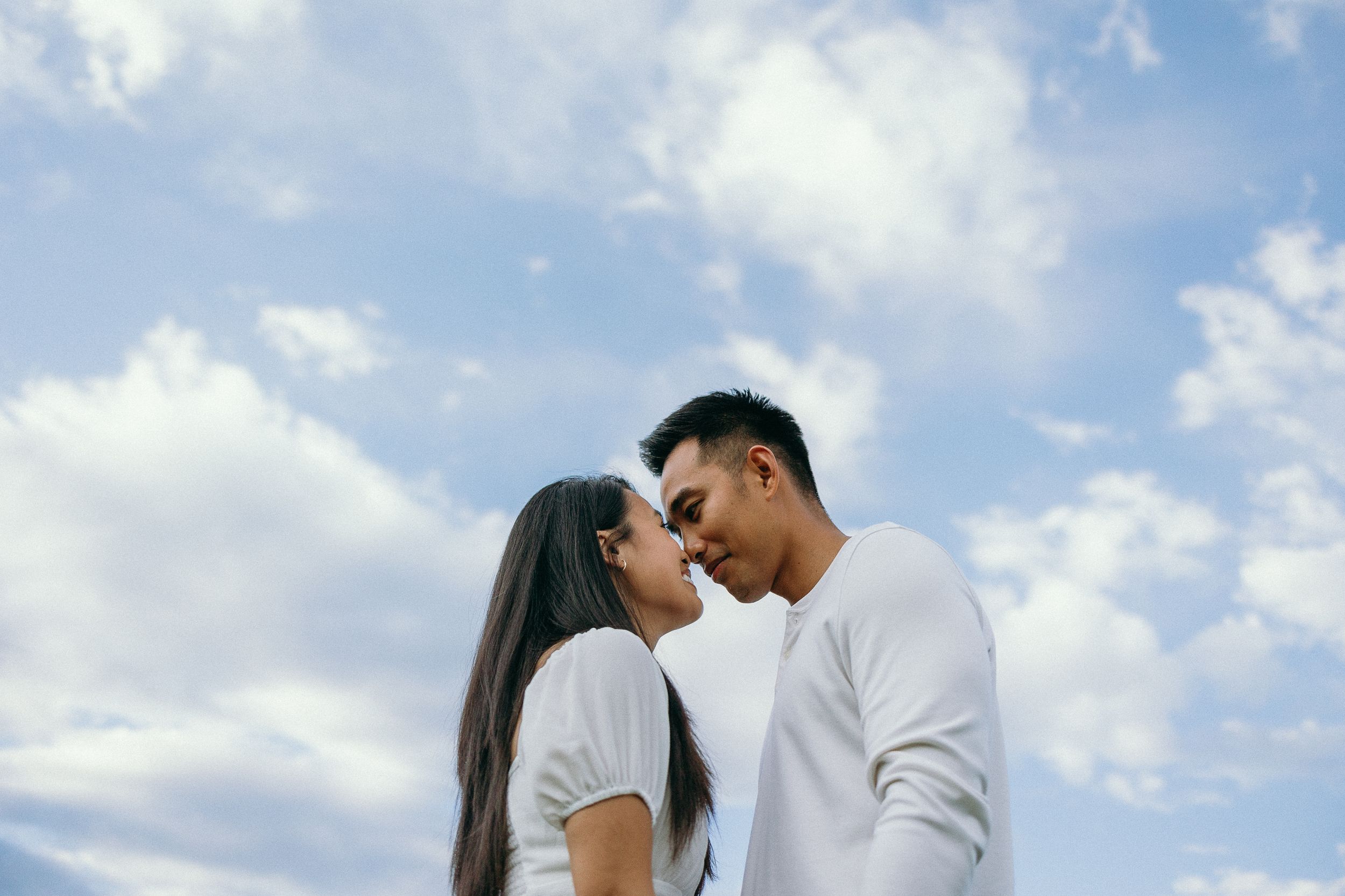 A couple shares an intimate moment in a sunny meadow with white clouds in the background.