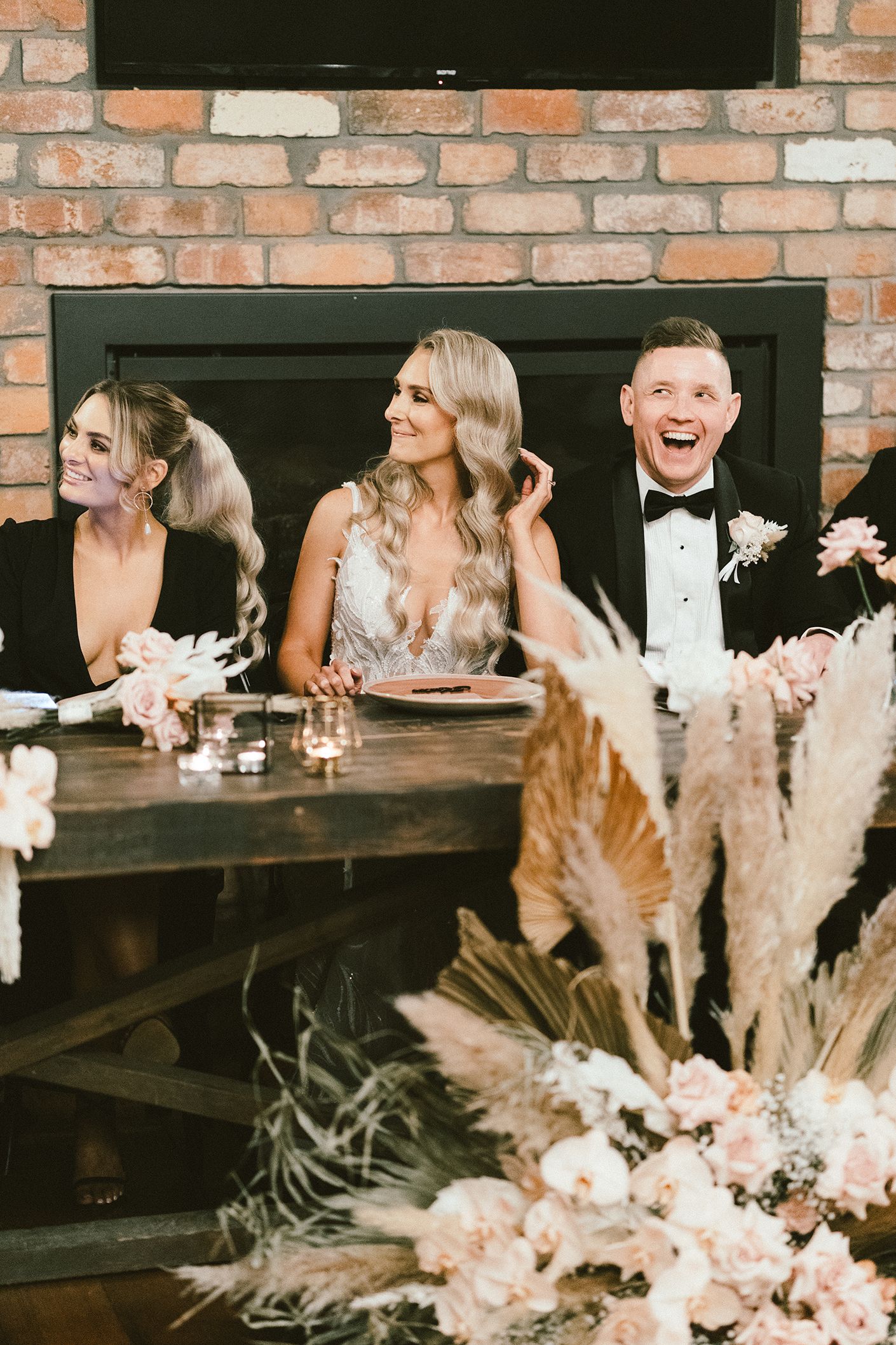 Wedding party seated at head table with rustic brick backdrop and pampas grass floral arrangements.