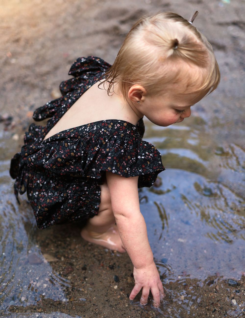 Small child in sparkly black dress exploring water puddle outdoors.
