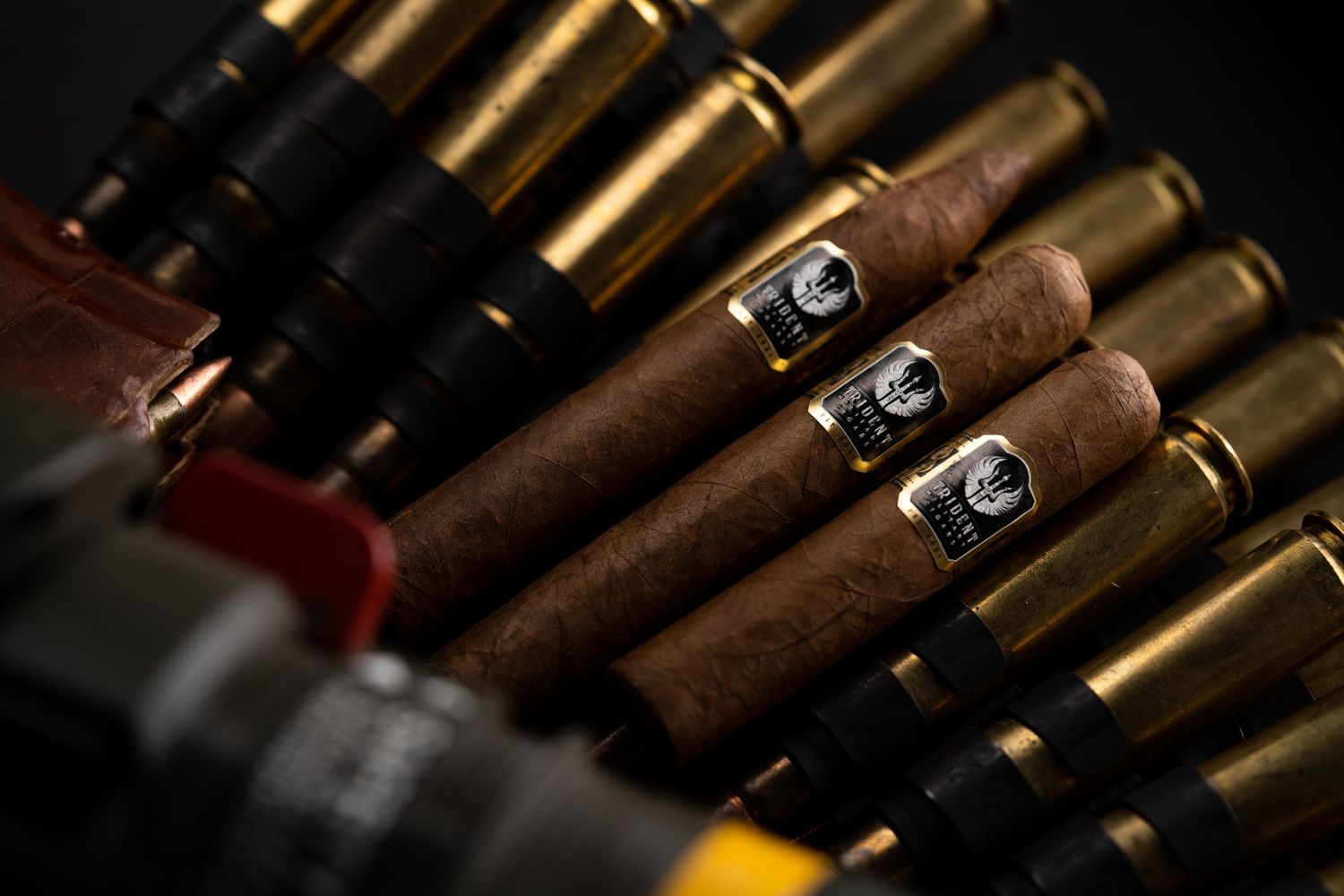 Close-up of cigars lined up alongside rifle ammunition against dark background.