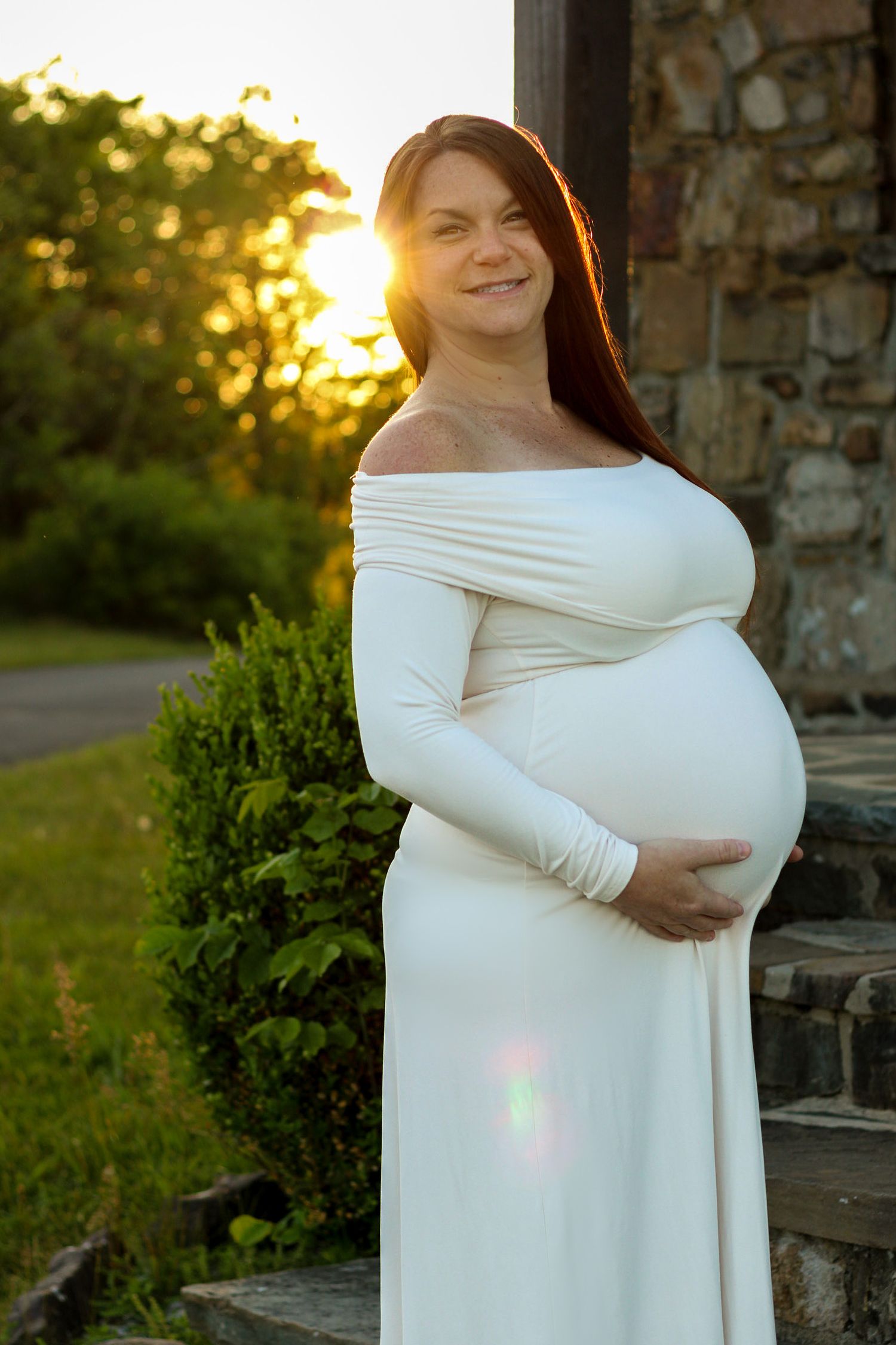 Maternity photo session at sunset with woman in flowing white off-shoulder dress.
