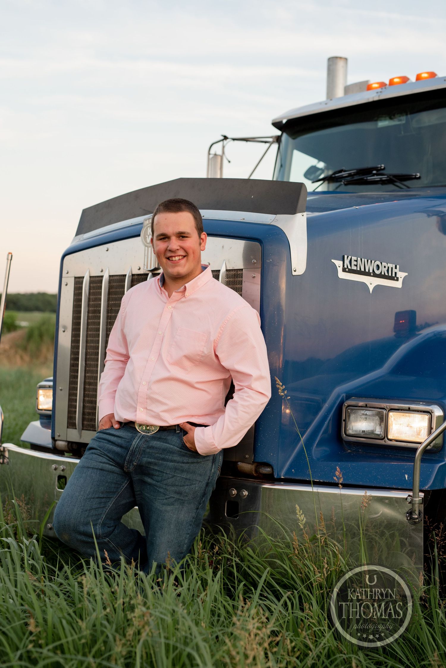 Professional portrait of driver in pink shirt leaning against blue Kenworth truck in grass.